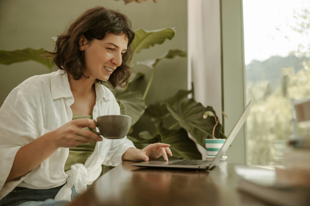 Smiling woman with a cup of tea at her laptop beside large green plants