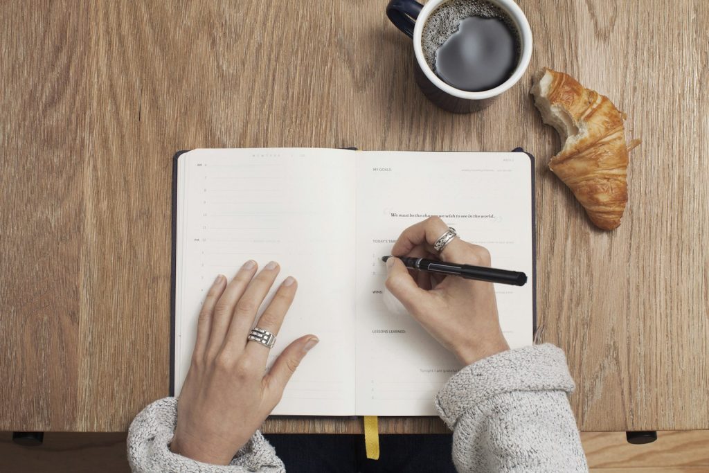 Hands writing in a journal beside a cup of coffee