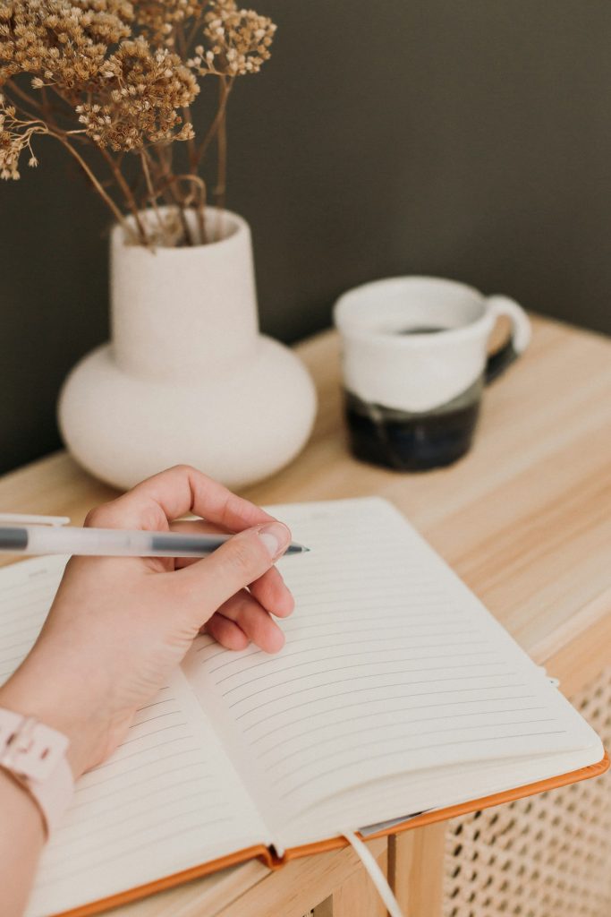 Hand writing in a notebook beside a vase and mug